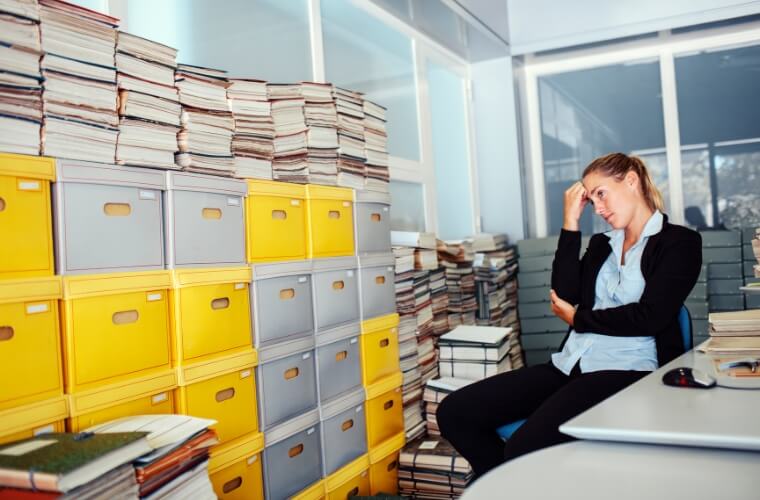 Woman looking distressed at messy office environment with archive of paper files