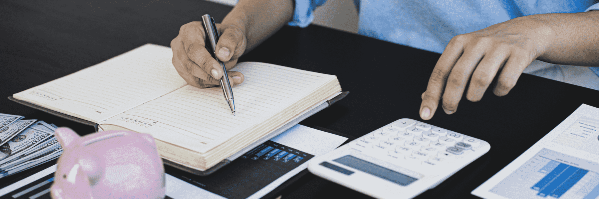 Person at a desk writing in a notebook with a calculator on the side